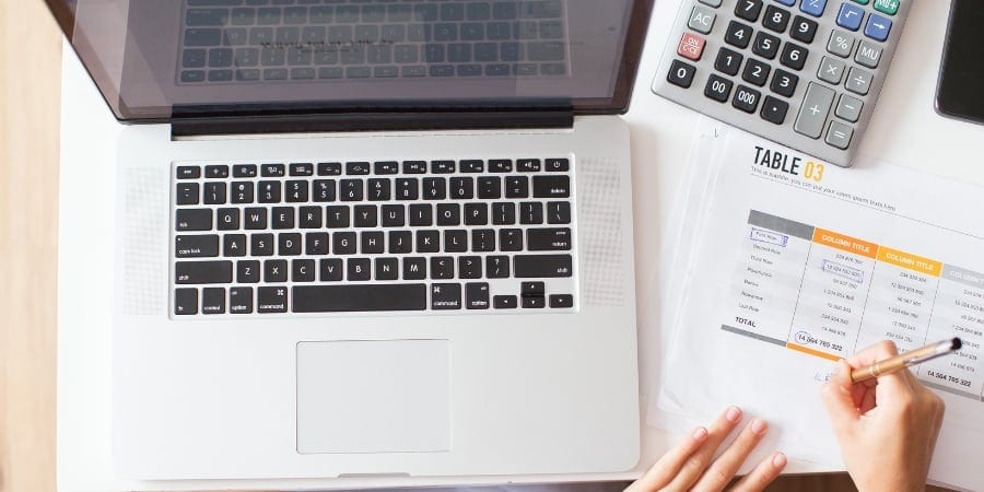 Mulher em frente a um computador com uma tabela de contas a pagar e a receber em mãos e fazendo contas para calculá-las